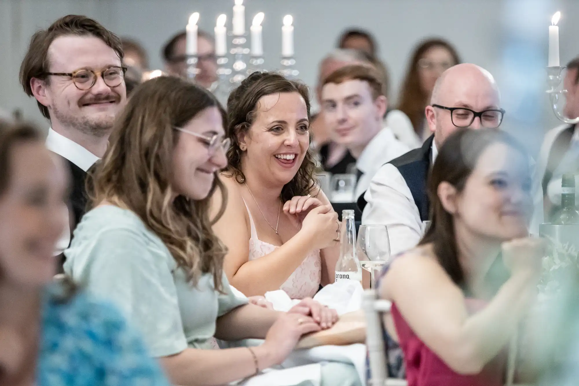 A group of people sitting at a formal event, with some smiling and others engaged. Candles and glassware are visible on the table.
