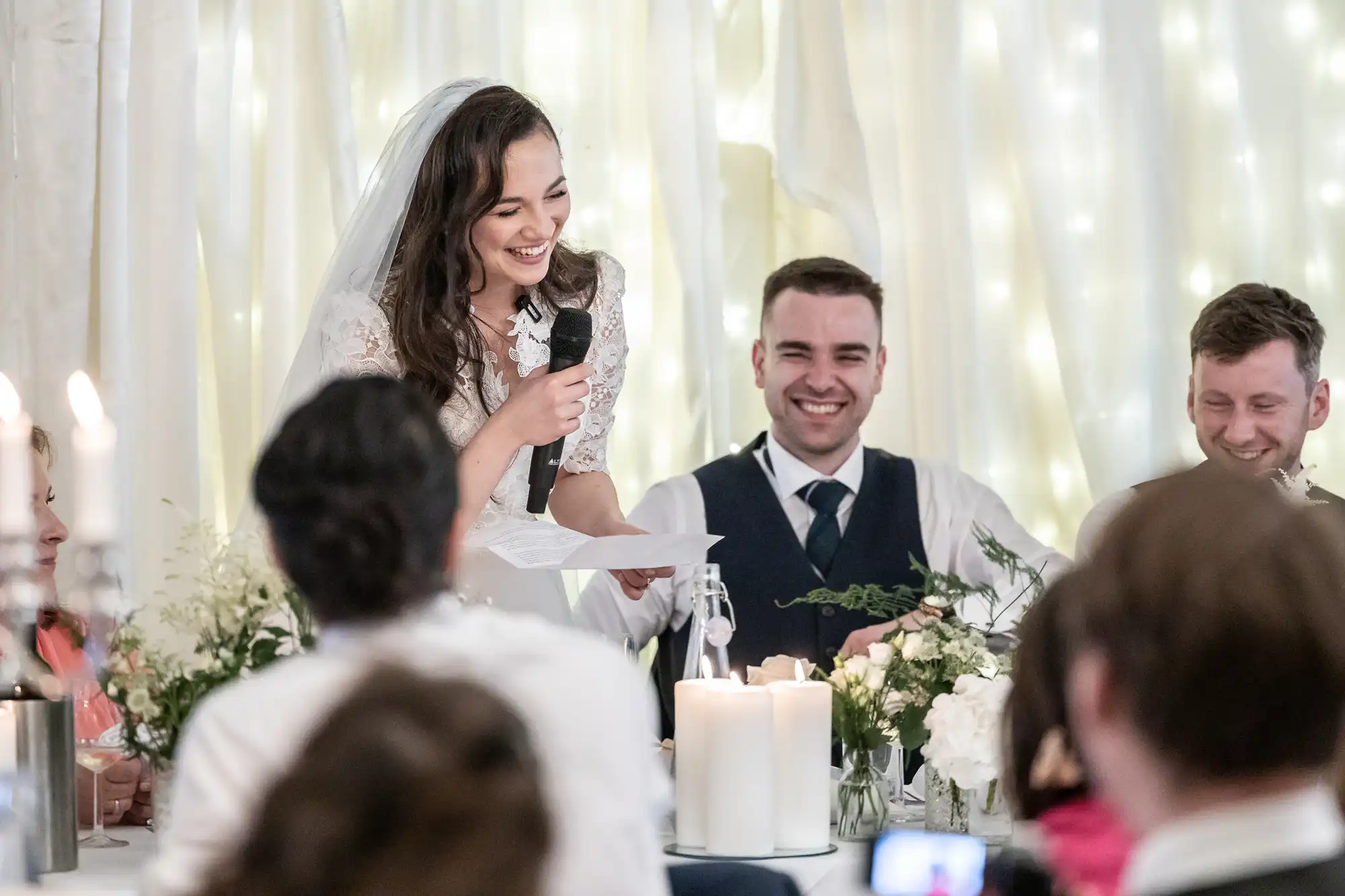 A bride smiles as she gives a speech with a microphone at a wedding reception, surrounded by people and decorations.