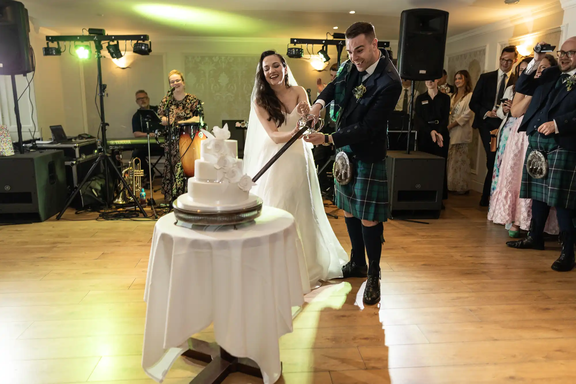 Bride and groom in kilts cutting a wedding cake with a sword in a reception hall, surrounded by guests and musical equipment.