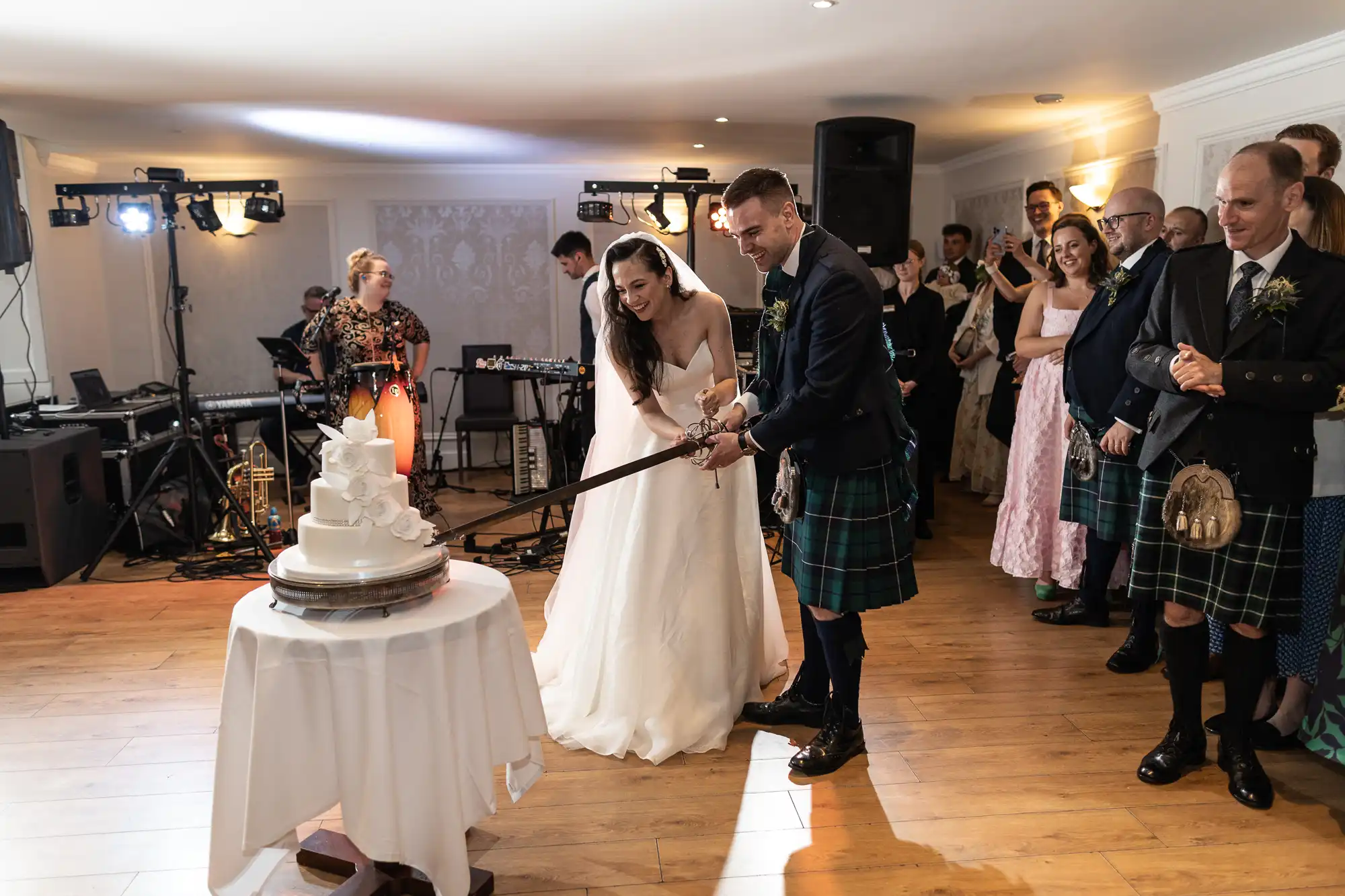 Bride and groom in formal attire cutting a tiered wedding cake with a sword, surrounded by guests at a reception.