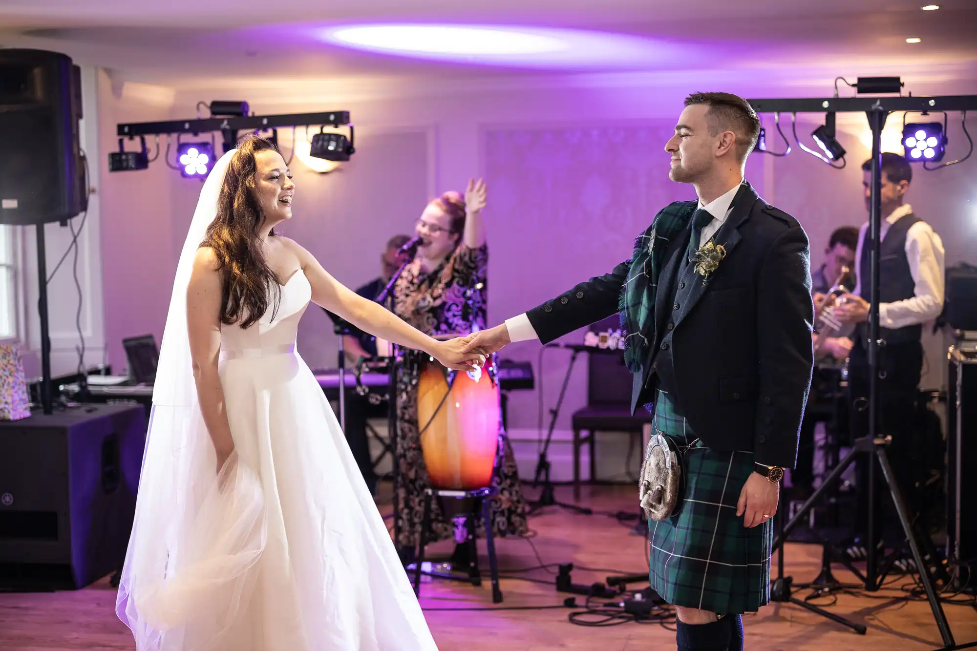 Bride and groom dance at a wedding reception, holding hands, with musicians performing in the background.
