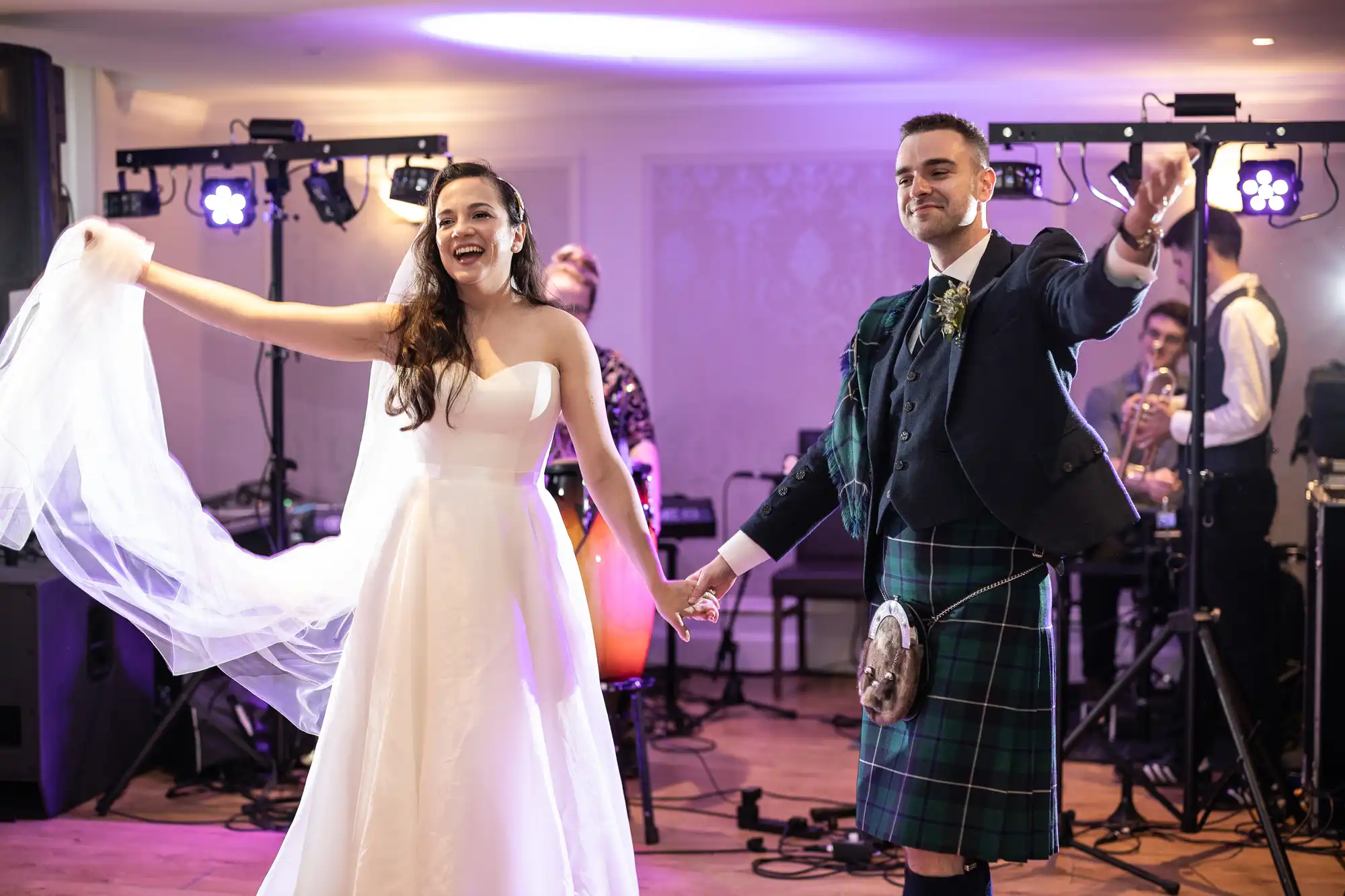 Bride and groom smile and hold hands during a wedding dance. The groom wears a kilt, and musicians play in the background.