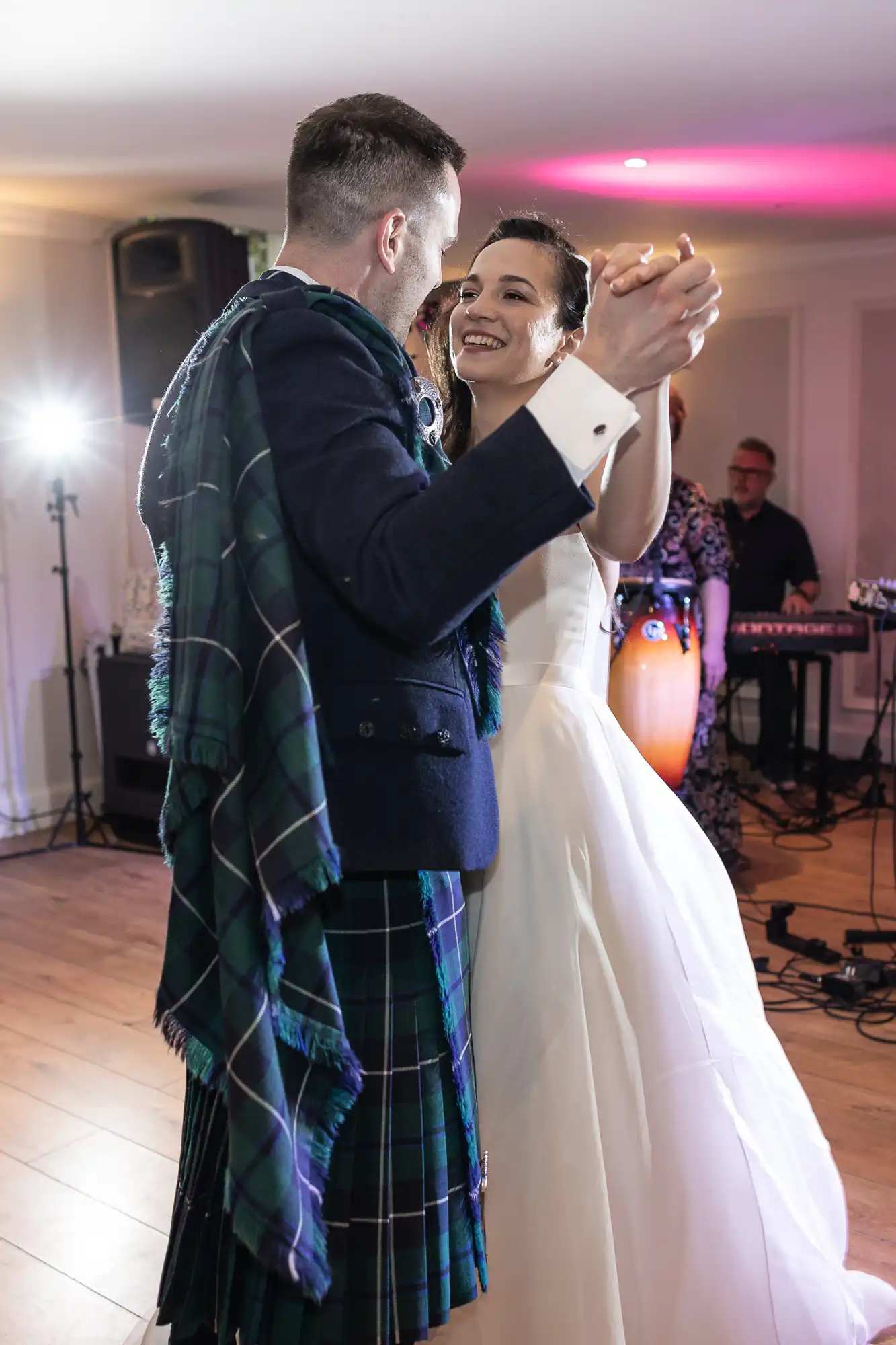 A bride and groom dance together in a reception hall. The groom is wearing a kilt, and there is a band playing in the background.