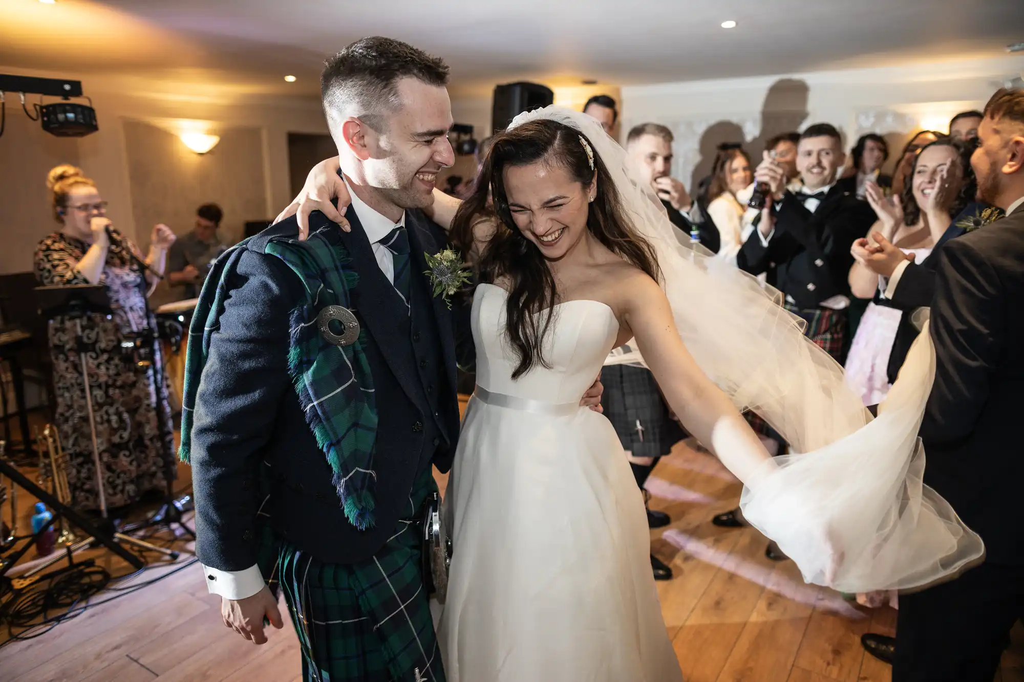 A bride and groom are smiling and dancing at their wedding reception. The groom is wearing a kilt, and there are guests clapping in the background.