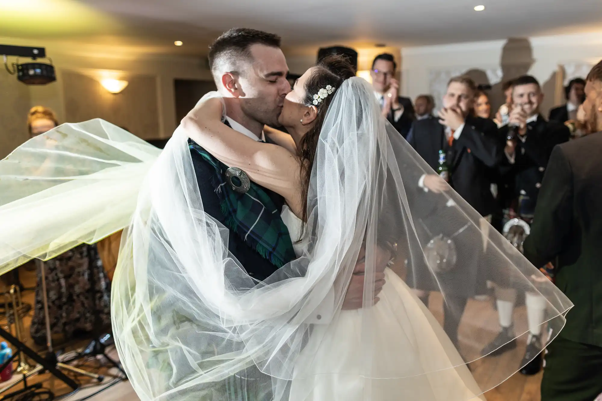 A bride and groom, wearing a white gown and a tartan outfit, kiss on a dance floor surrounded by guests with some clapping and smiling.