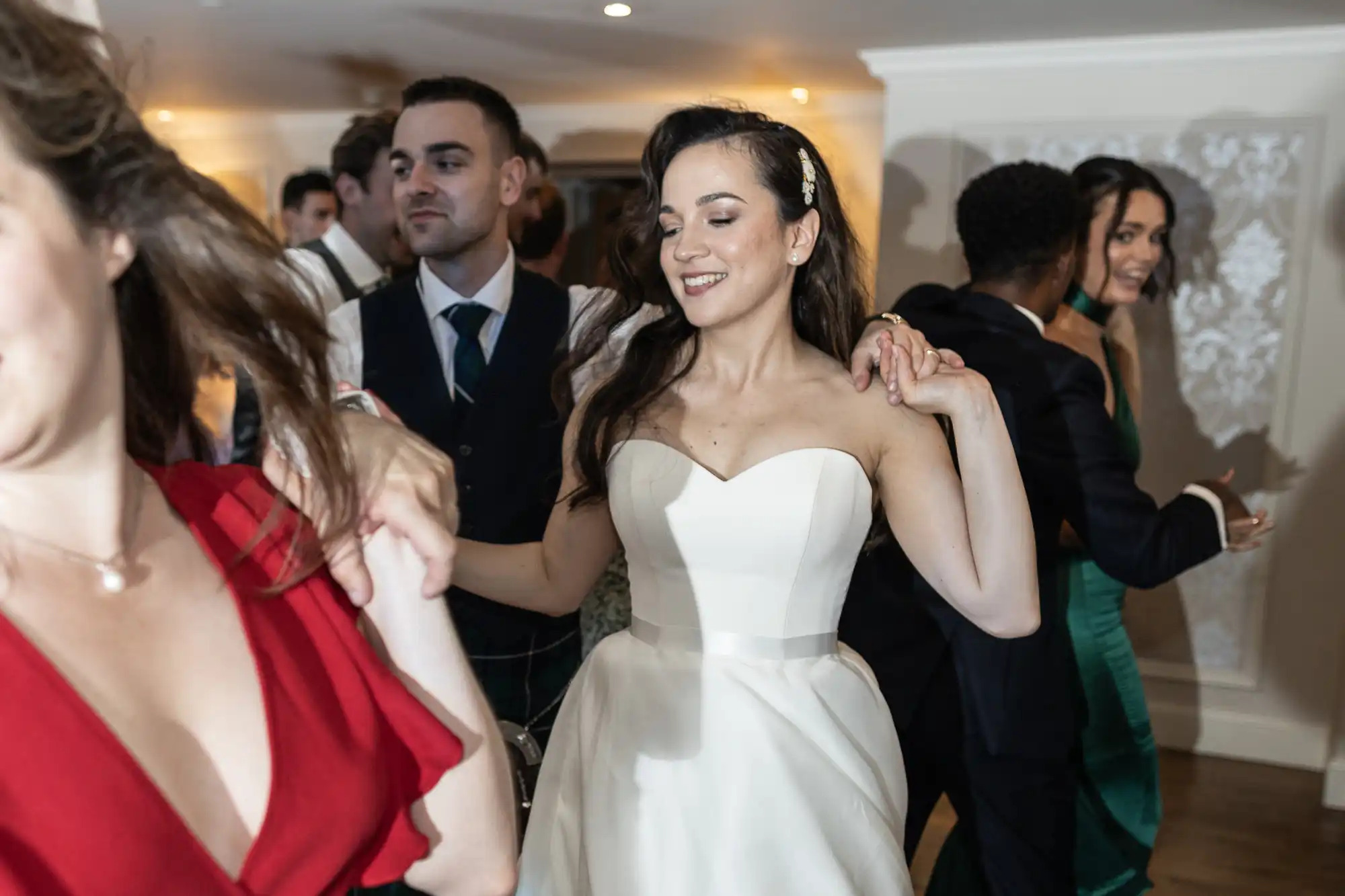 A bride in a white dress dances joyfully with guests at a lively indoor wedding reception.