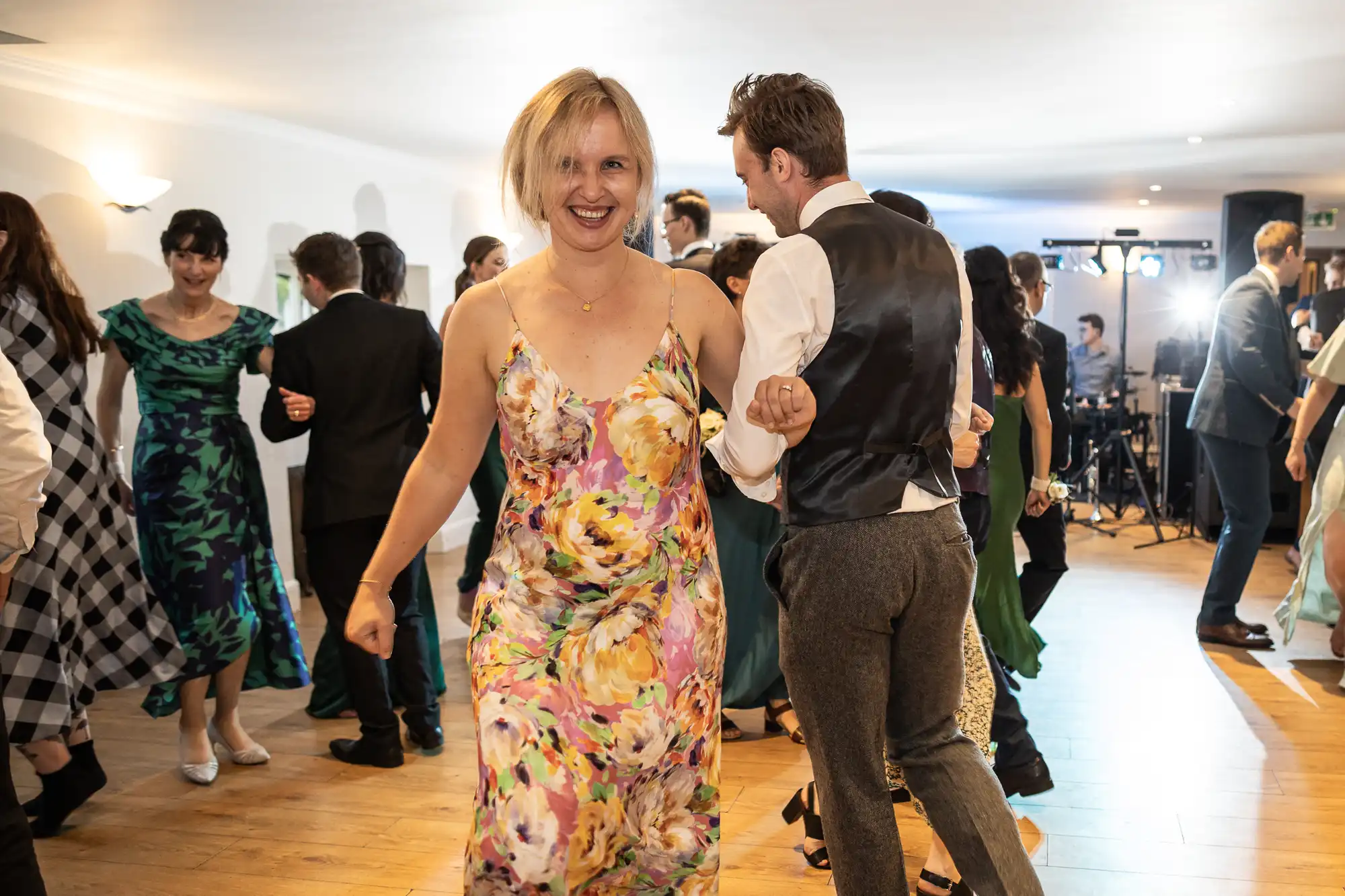 A woman in a floral dress smiles while dancing at an indoor event surrounded by other people in formal attire.