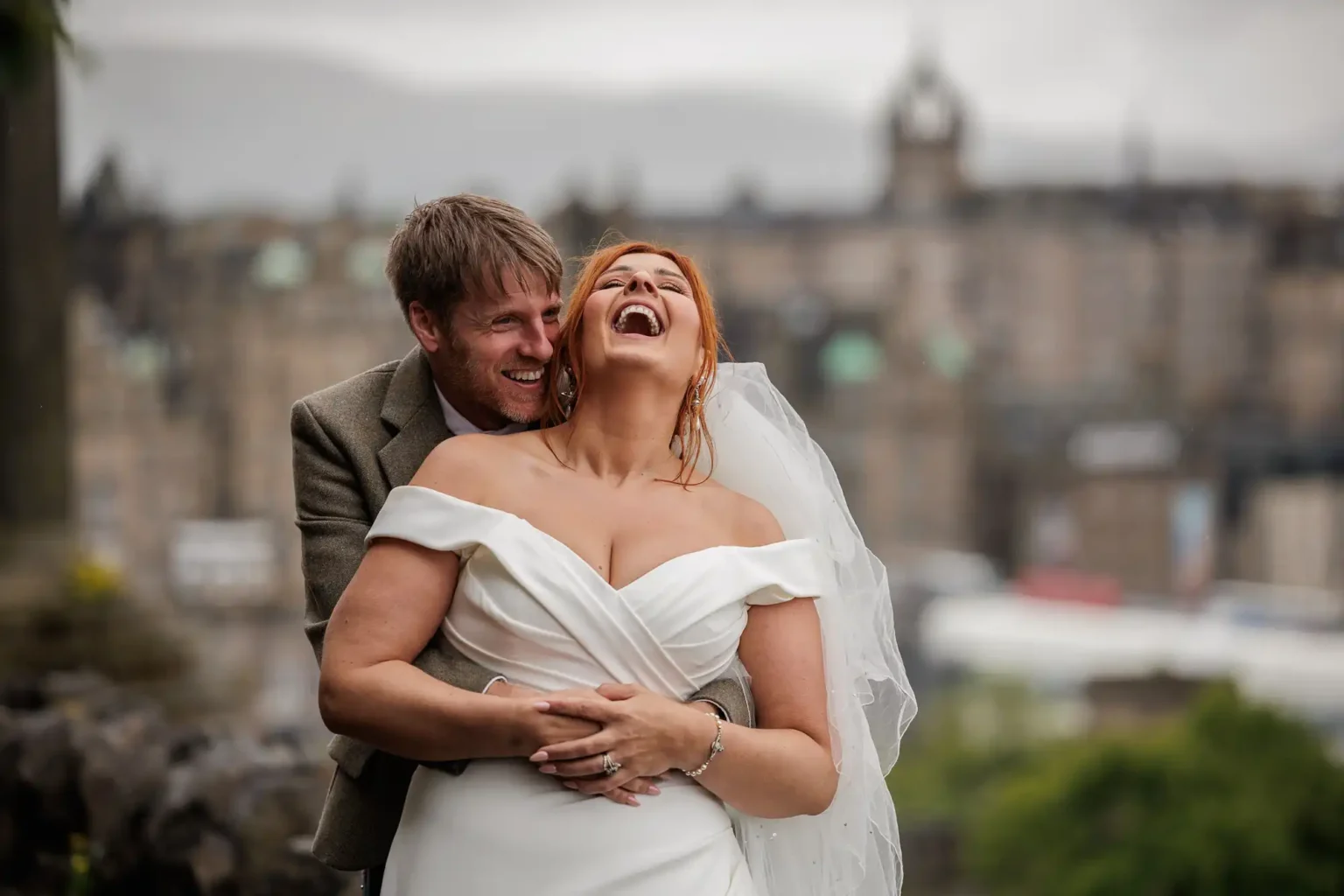 A bride in a white dress and veil laughs as a groom in a suit embraces her from behind outdoors, with blurred city buildings in the background at Calton Hill, Edinburgh, created as Edinburgh wedding videographer.