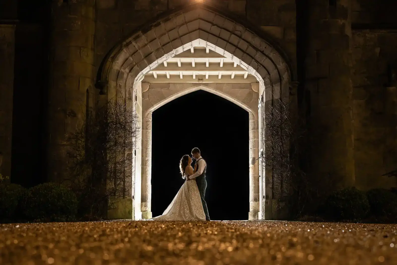 A bride and groom stand embraced under a large stone archway at night, illuminated by lights from behind by Dundas Castle, Edinburgh wedding photographers.