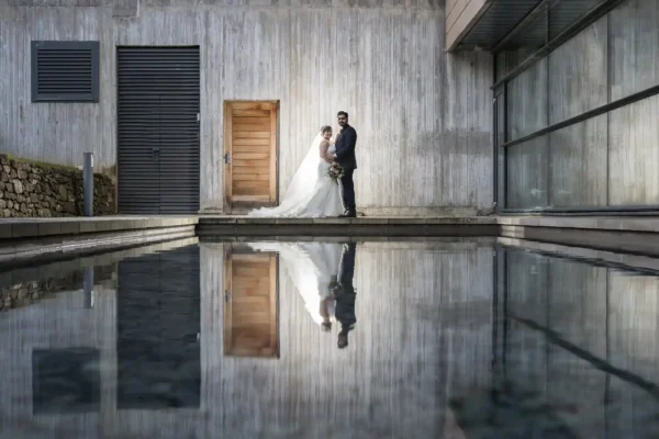 A bride in a white dress and groom in a dark suit stand close together by a wooden door against a modern concrete wall, their reflection visible in a calm rectangular pool in the foreground. Light highlights the couple, contrasting with the grey background.