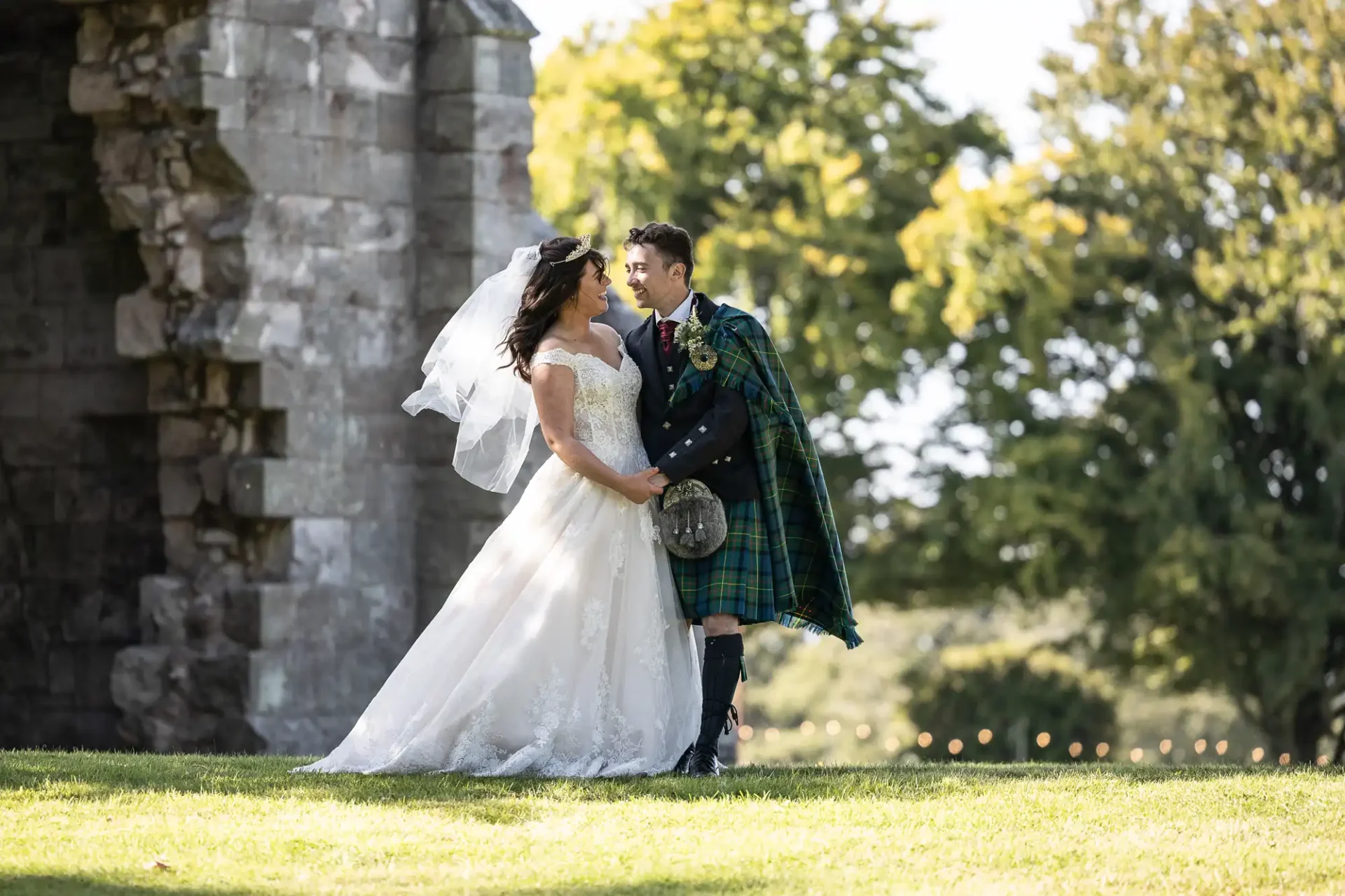 Bride and groom standing outdoors, holding hands and smiling at each other, captured by their Dunglass Estate wedding photographer. Groom wears a kilt and tartan attire; bride wears a white gown. Dunglass Church and trees in the background.