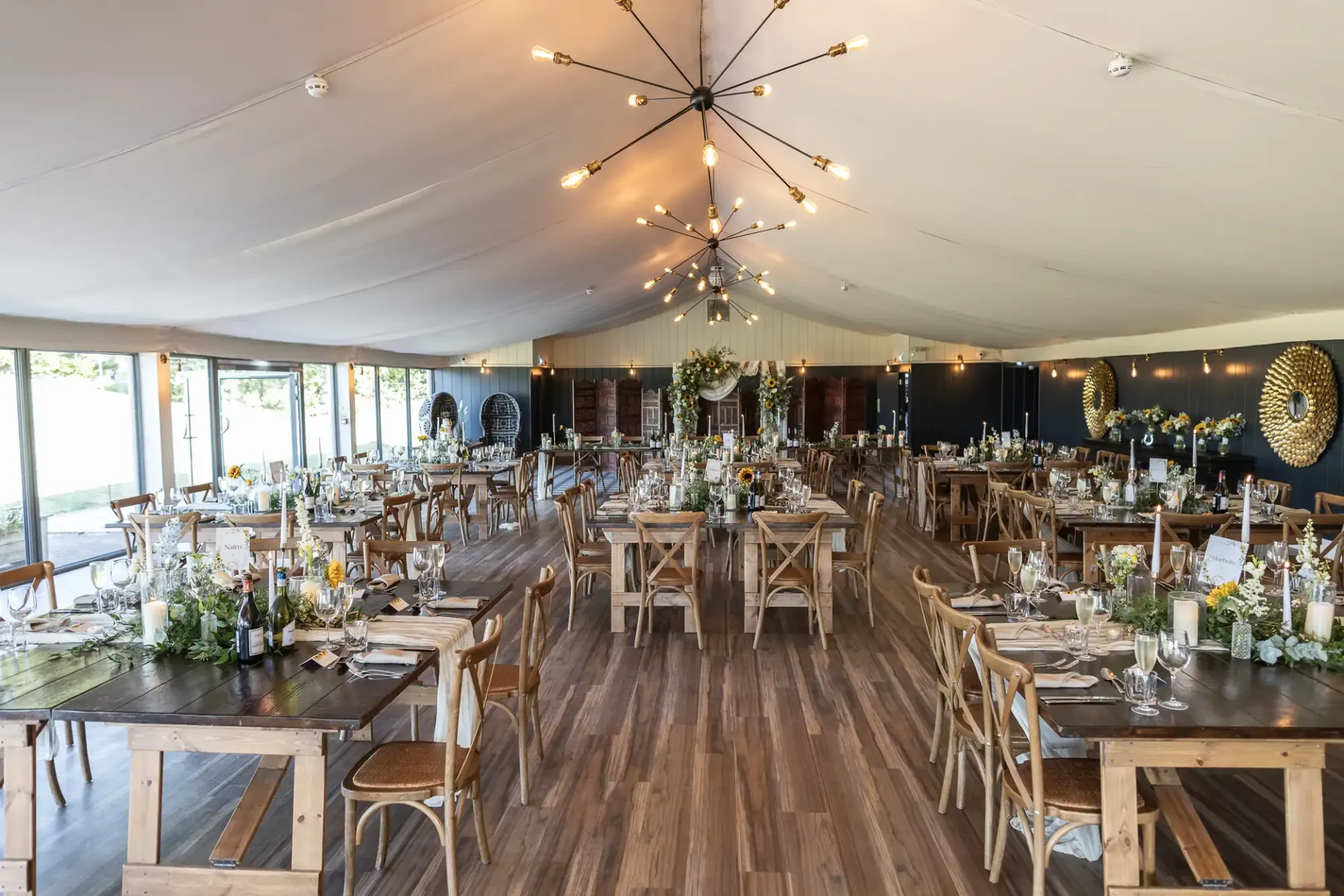 Spacious event hall, Dunglass Estate Pavilion, with wooden flooring, set with rows of wooden tables and chairs. Chandeliers hang from the ceiling, and floral decorations adorn the tables. Large windows are on one side.