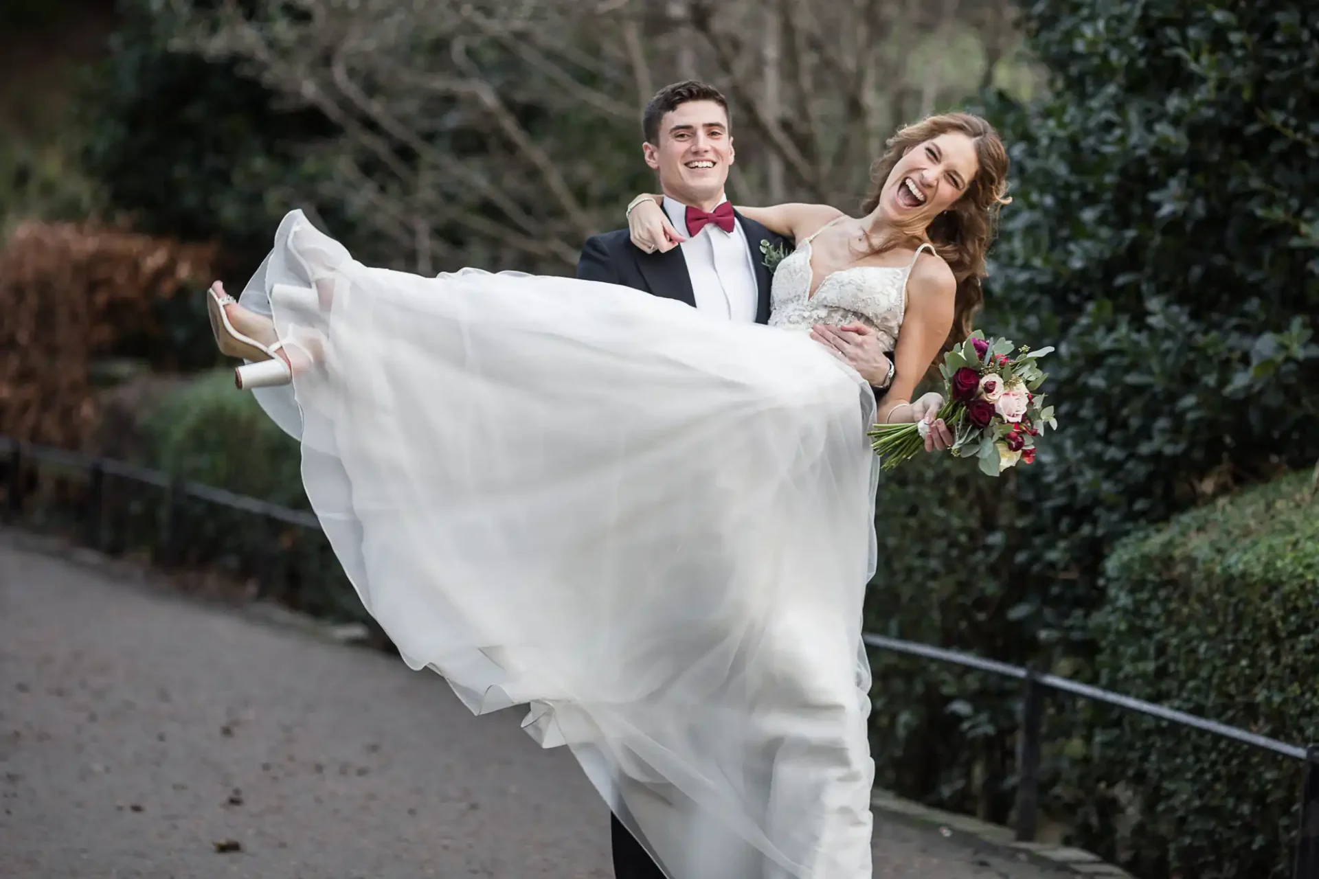 A groom in a black suit holds a smiling bride in a white wedding dress and holding a bouquet, outdoors on a pathway at Princes Street Gardens, Edinburgh, by wedding photographers in Edinburgh.