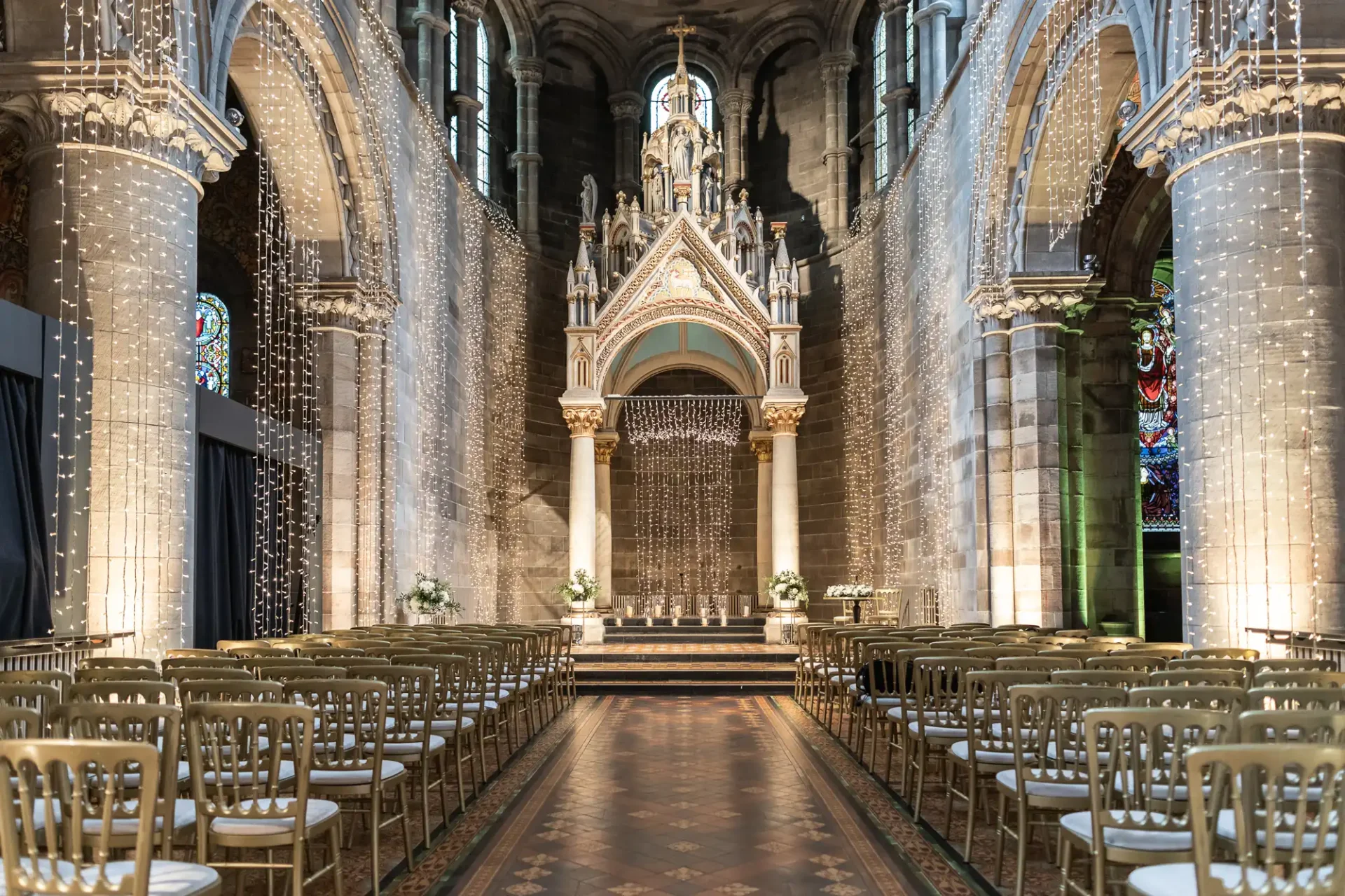 The Nave and Baldacchino at Mansfield Traquair; Ornate church interior set for a wedding, with rows of gold chairs, decorative arches, stained glass windows, and string lights.