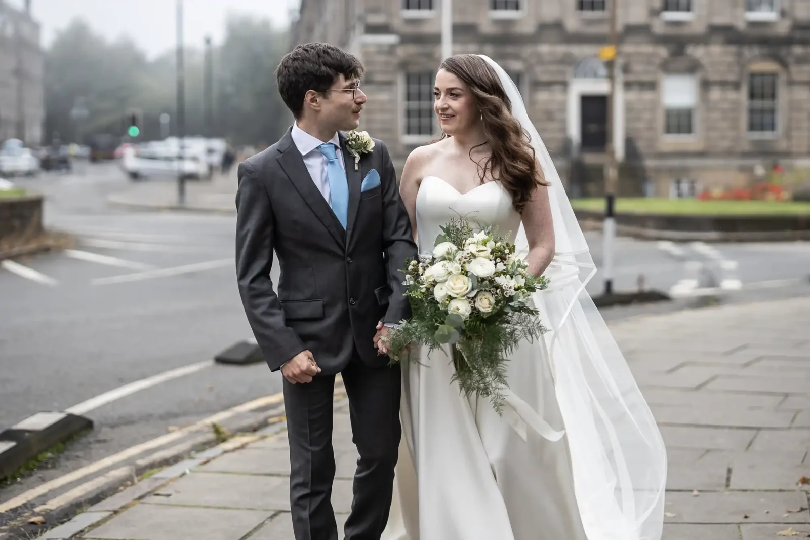 A bride and groom in formal attire walk hand in hand on an urban sidewalk, surrounded by buildings. The bride holds a bouquet of white flowers at Mansfield Traquair Edinburgh.