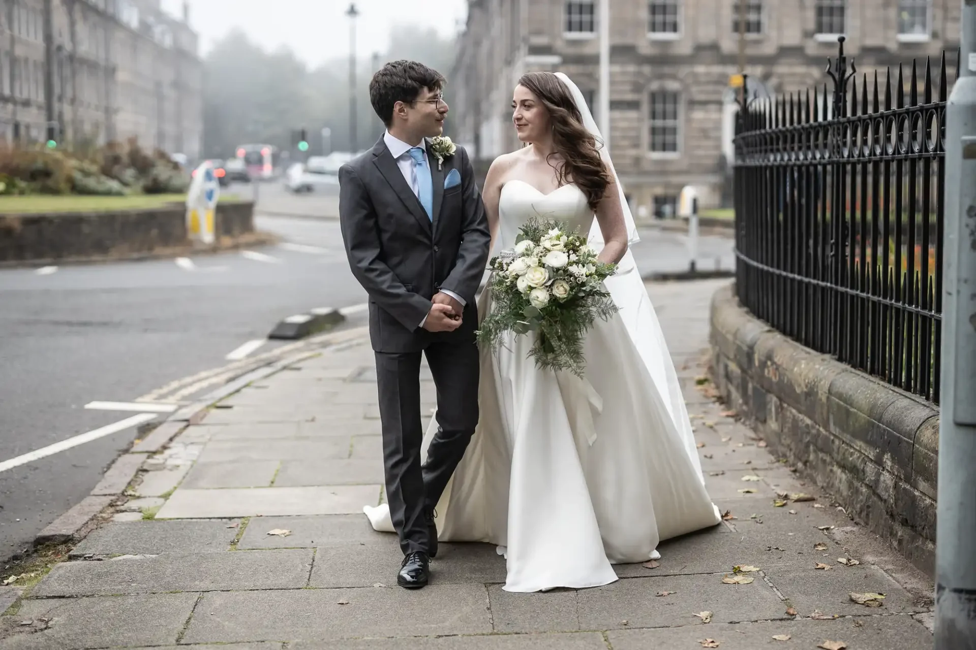 A bride in a white gown walks with a groom in a gray suit on a sidewalk. She holds a bouquet of white flowers. Buildings and a street are in the background at Mansfield Place, Edinburgh.