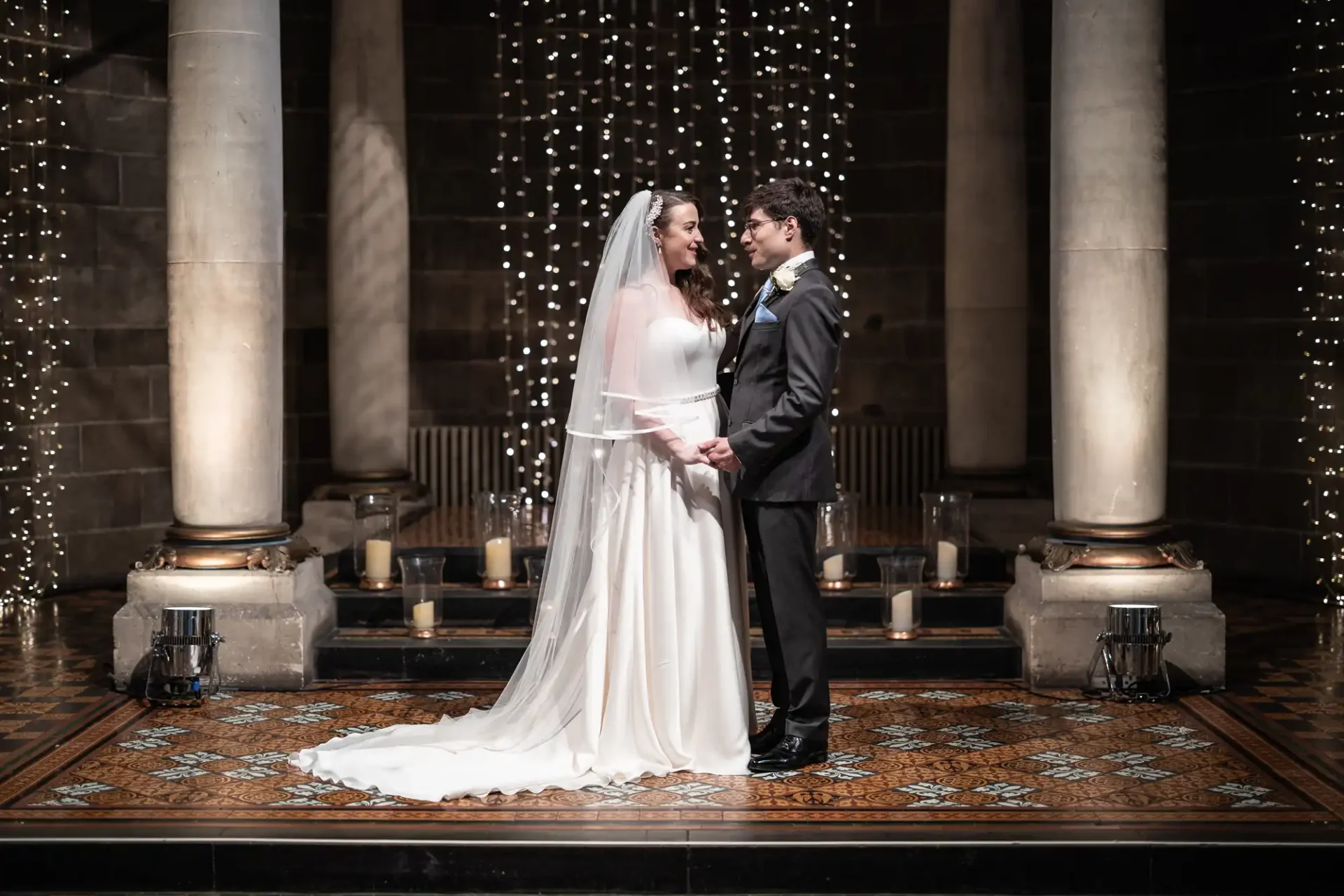 Bride and groom stand facing each other, holding hands, in a decorated venue, The Baldacchino at Mansfield Traquair, with columns and string lights.