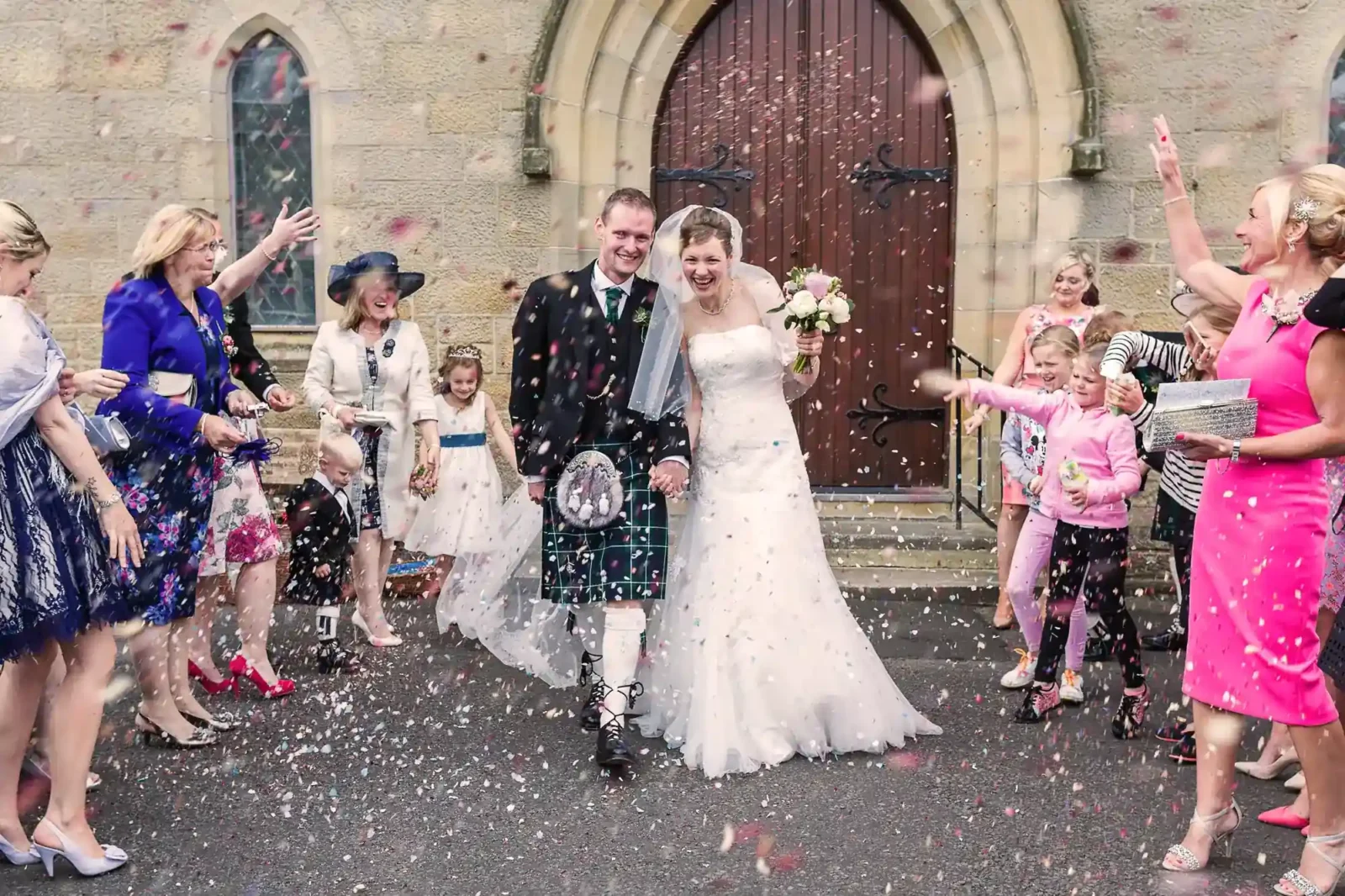A bride and groom, the groom in a kilt, exit a church smiling as guests shower them with confetti. Children and adults cheer, some dressed in bright colours, as the couple hold hands and walk past a stone arch doorway. The mood is joyful and festive.