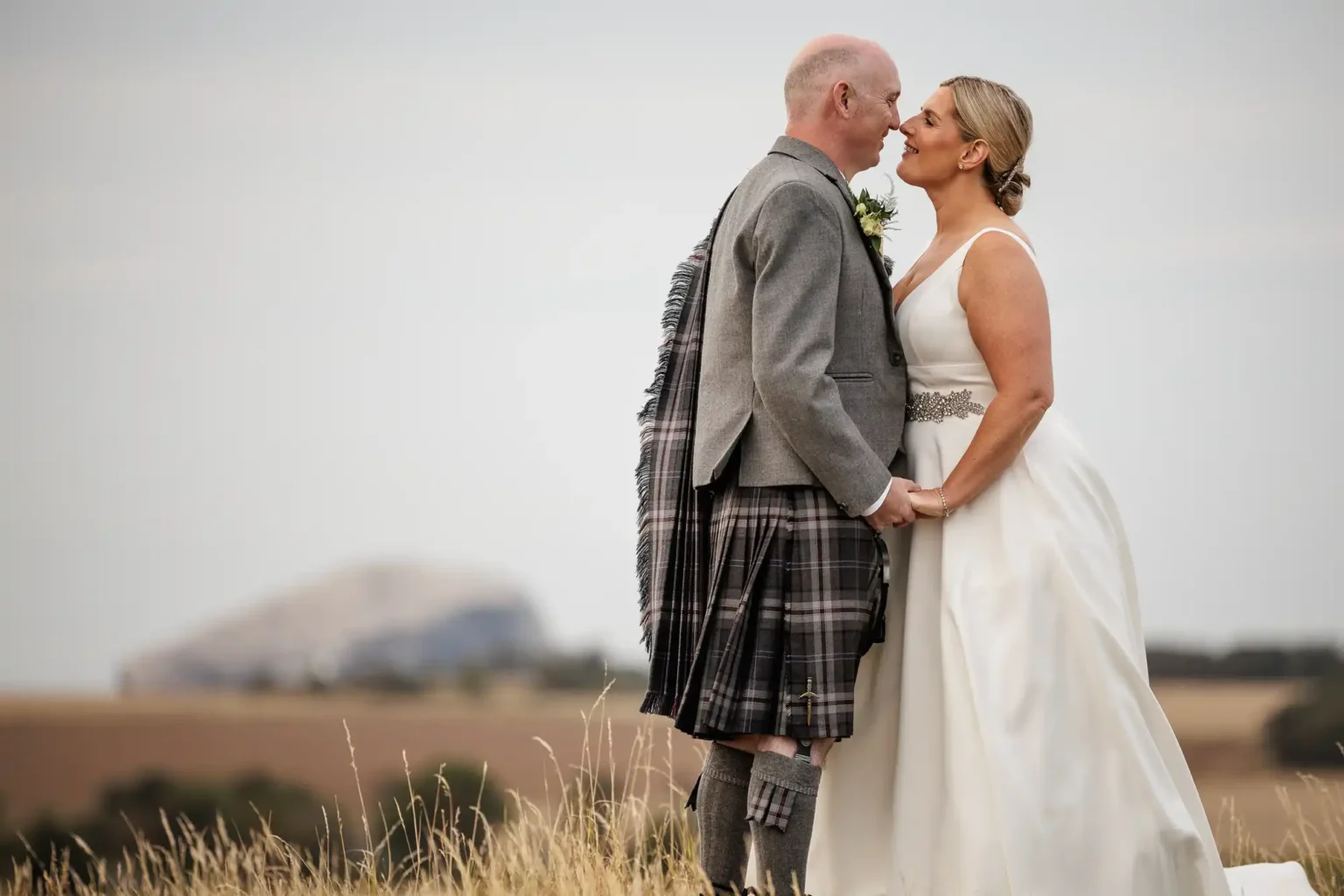 Whitekirk Hill wedding photographer photo of a couple stands in a field holding hands, both smiling and touching noses. The groom wears a grey jacket and checked kilt, while the bride wears a white sleeveless dress. Distant hills and a large rocky formation are visible in the background under a cloudy sky.