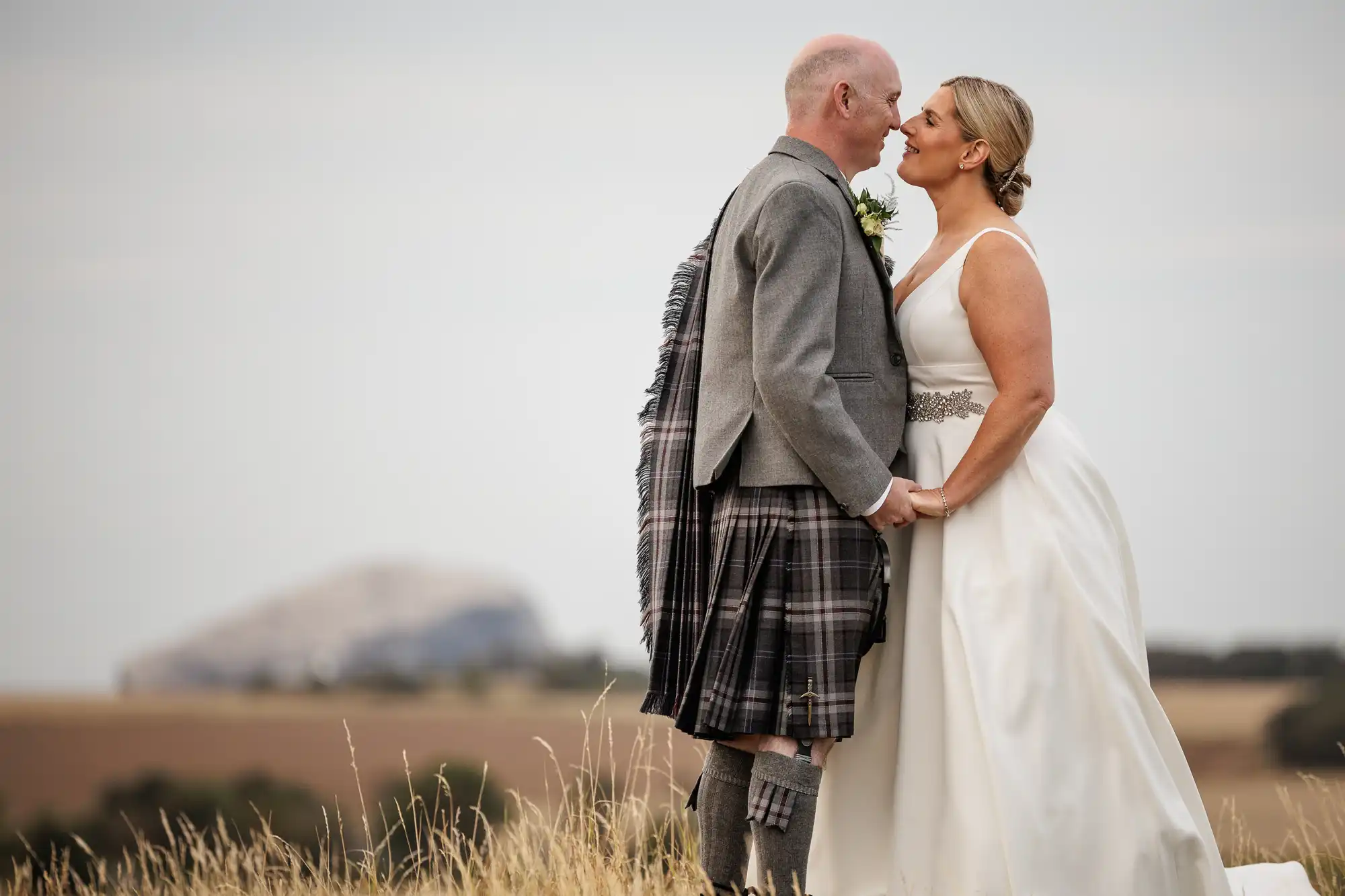 Whitekirk Hill wedding photographer photo of a couple stands in a field holding hands, both smiling and touching noses. The groom wears a grey jacket and checked kilt, while the bride wears a white sleeveless dress. Distant hills and a large rocky formation are visible in the background under a cloudy sky.