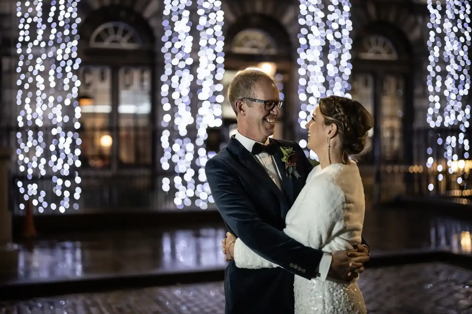 A couple in wedding attire embrace and smile at each other outdoors at night. Behind them, tall windows and hanging strings of white lights create a festive atmosphere. The ground is wet, reflecting the lights. Both appear joyful and happy.