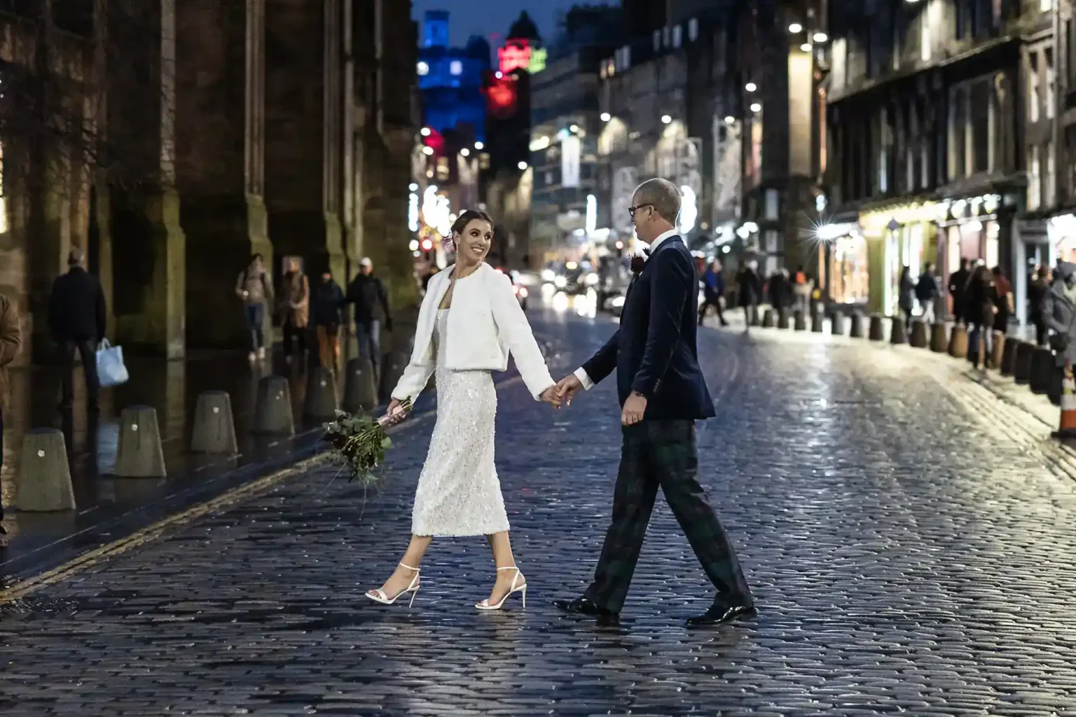 A couple in wedding attire smile and hold hands while walking across a cobbled street at night, city lights and people in the background. The woman carries a bouquet and wears white; both look joyful.