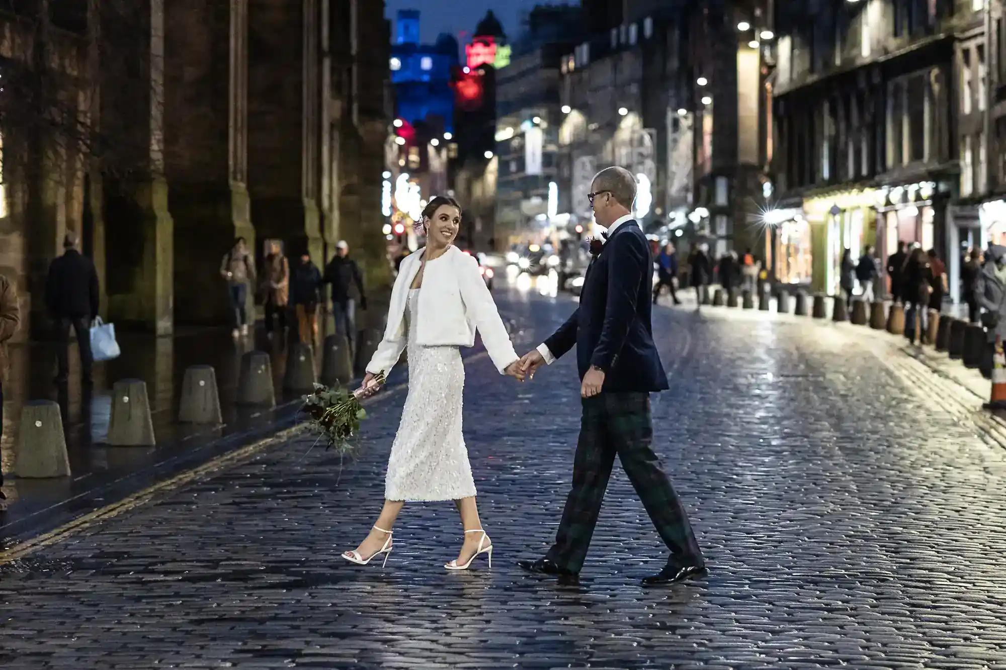 A couple in wedding attire smile and hold hands while walking across a cobbled street at night, city lights and people in the background. The woman carries a bouquet and wears white; both look joyful.